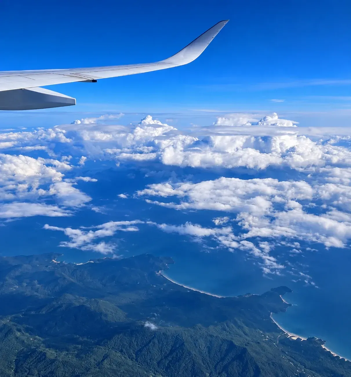 Asa de avião sobre nuvens brancas e o litoral verde visto do alto