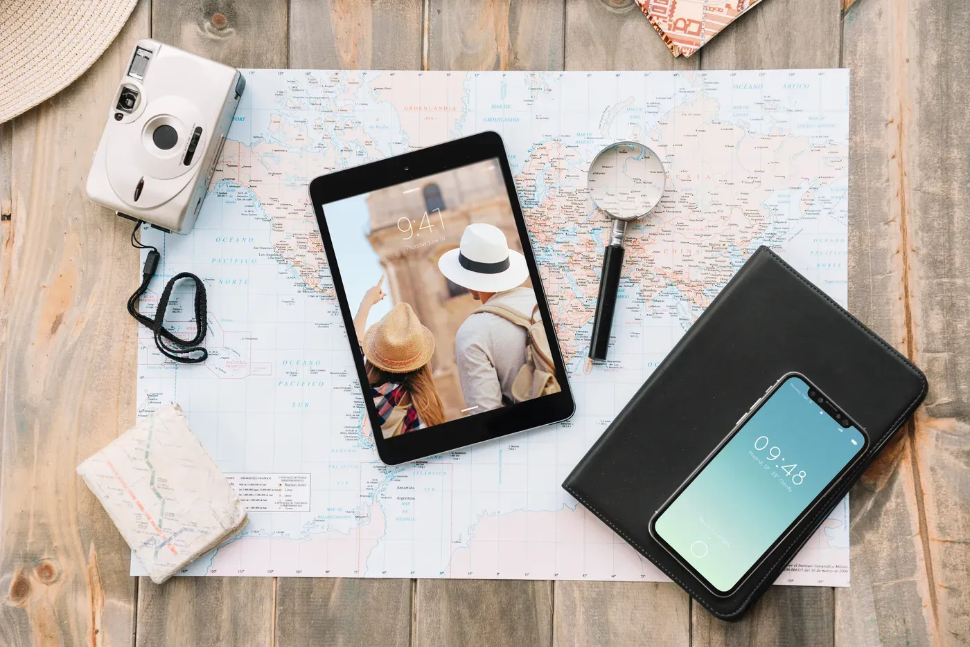 Mesa de madeira com mapa-múndi, tablet, câmera, lupa e celular usados no planejamento de um roteiro de viagem